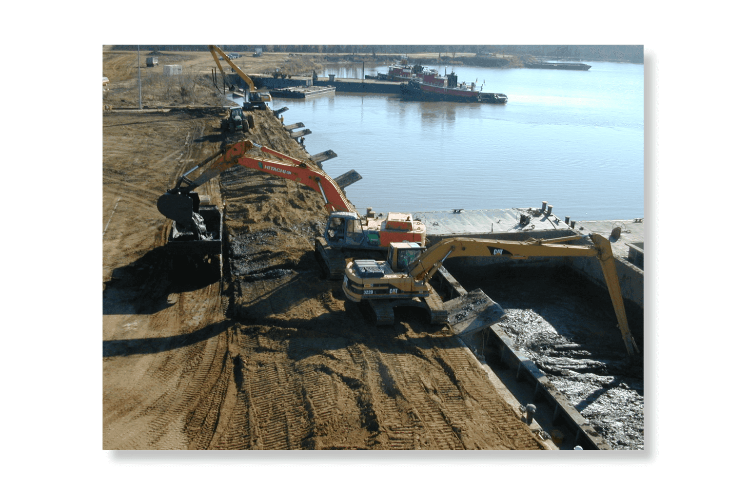 An excavator working dredged material on a barge alongside the bulkhead, with the river and moored vessels behind.