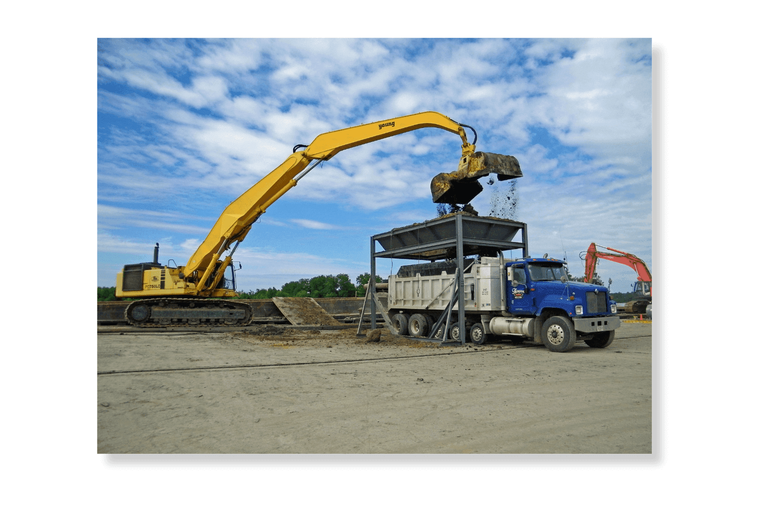 A long-reach excavator loading dredged material into a dump truck at a laydown area under a blue sky.