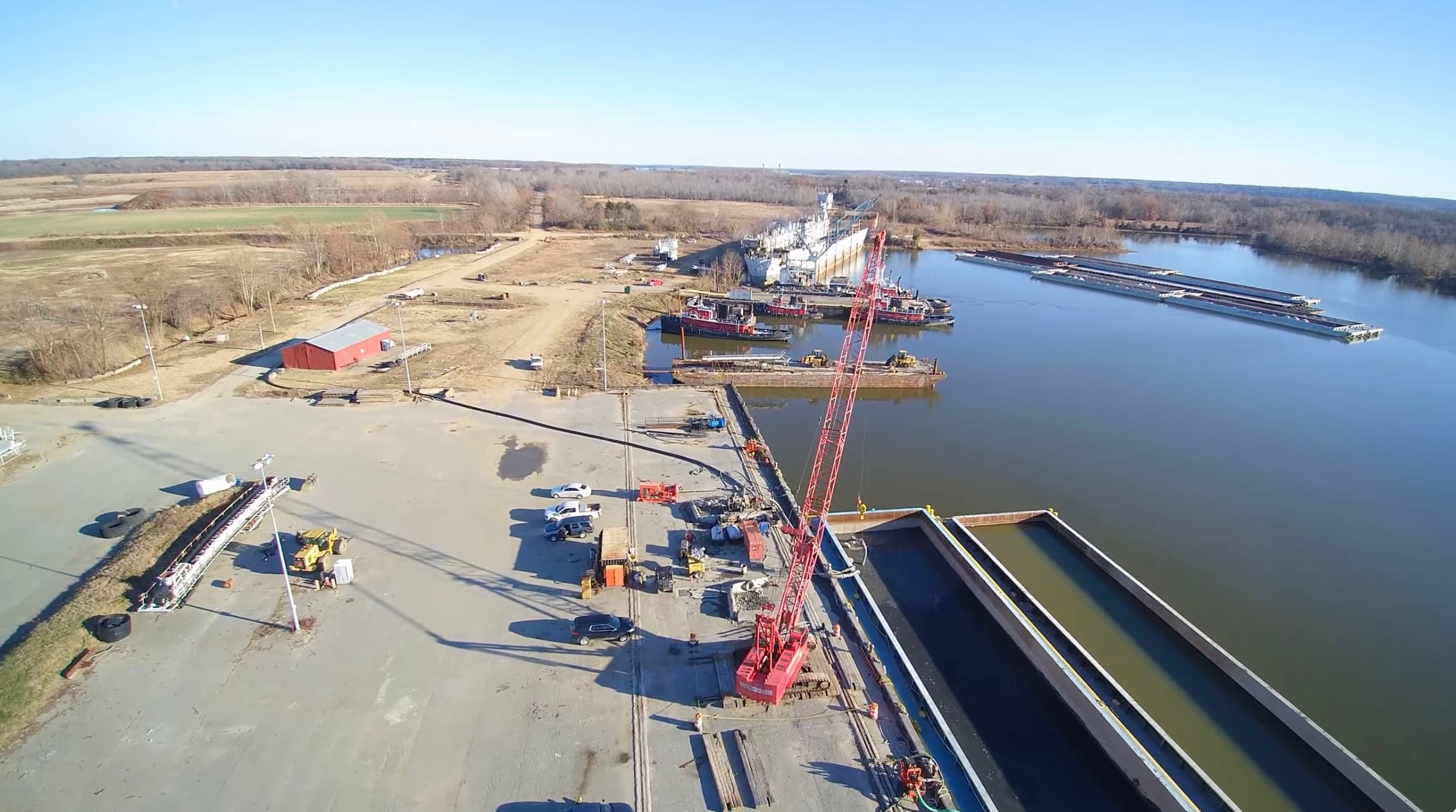 Aerial view of Weanack Land's Port Tobacco terminal on the James River, showing the laydown yard, red crawler crane, red barn, moored tugboats, a ship, a tow of barges, and containment basins in the foreground