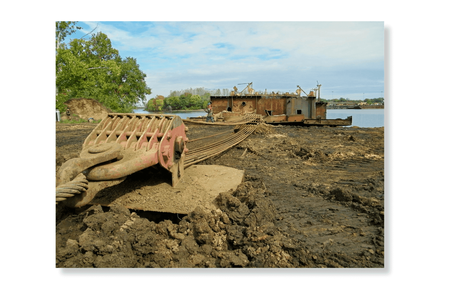A dredge cutterhead resting on the shoreline at Port Tobacco with a barge moored on the James River behind it.
