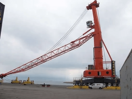 An assembled orange mobile harbor crane standing upright on its multi-wheeled undercarriage on a paved dock, its red lattice boom luffed down to a low resting angle. A white van parked at the base of the crane gives a sense of scale, with calm water and an overcast sky behind.